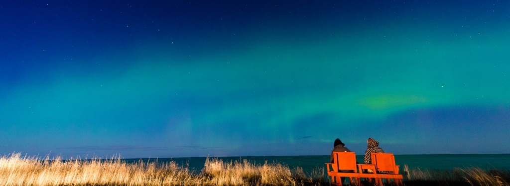 View over Lake Superior at night with Northern Lights in the clouds above; two people are sitting in chairs watching.