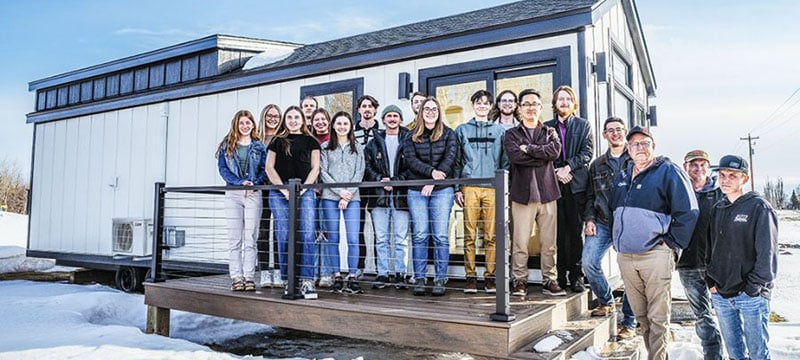 17 people stand on the porch of a model mini home by Brockway Mini Homes including a dozen students from Michigan Tech’s marketing capstone class.