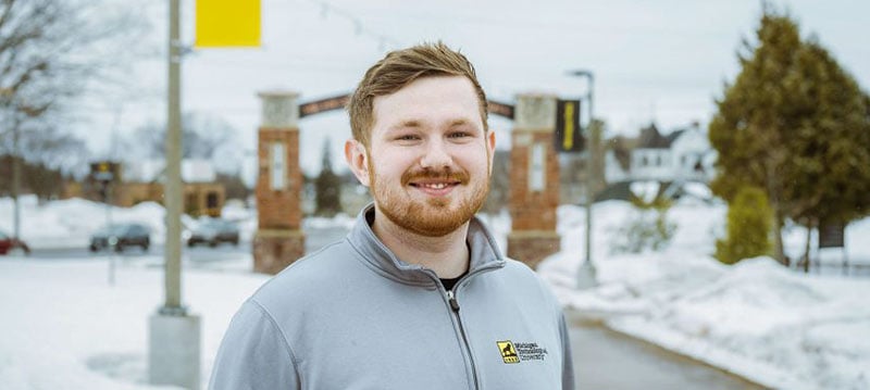 A student wearing a Michigan Technological University shirt stands under the Alumni Gateway Arch at Michigan Tech as he prepares to collect his second degree from the institution.