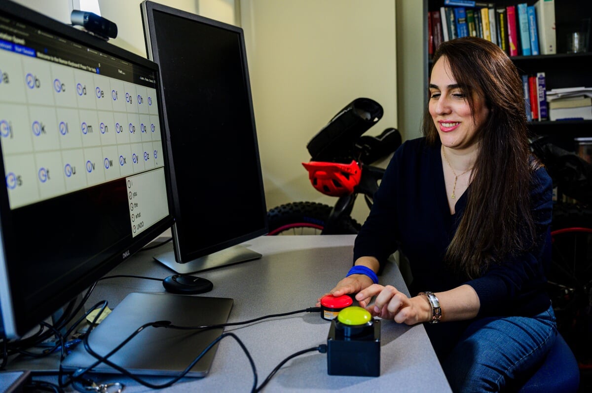 Graduate student pushes a red and a neon-yellow button with characters on the computer screen in front of her in an office setting.