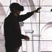Person wearing VR goggles reaching out toward a building on a screen.