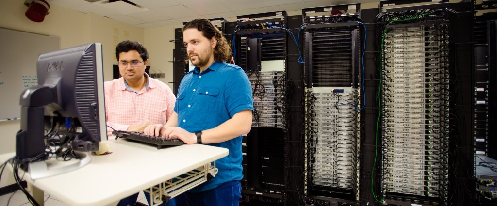 Two graduate students standing in front of a computer with a wall of servers behind them.