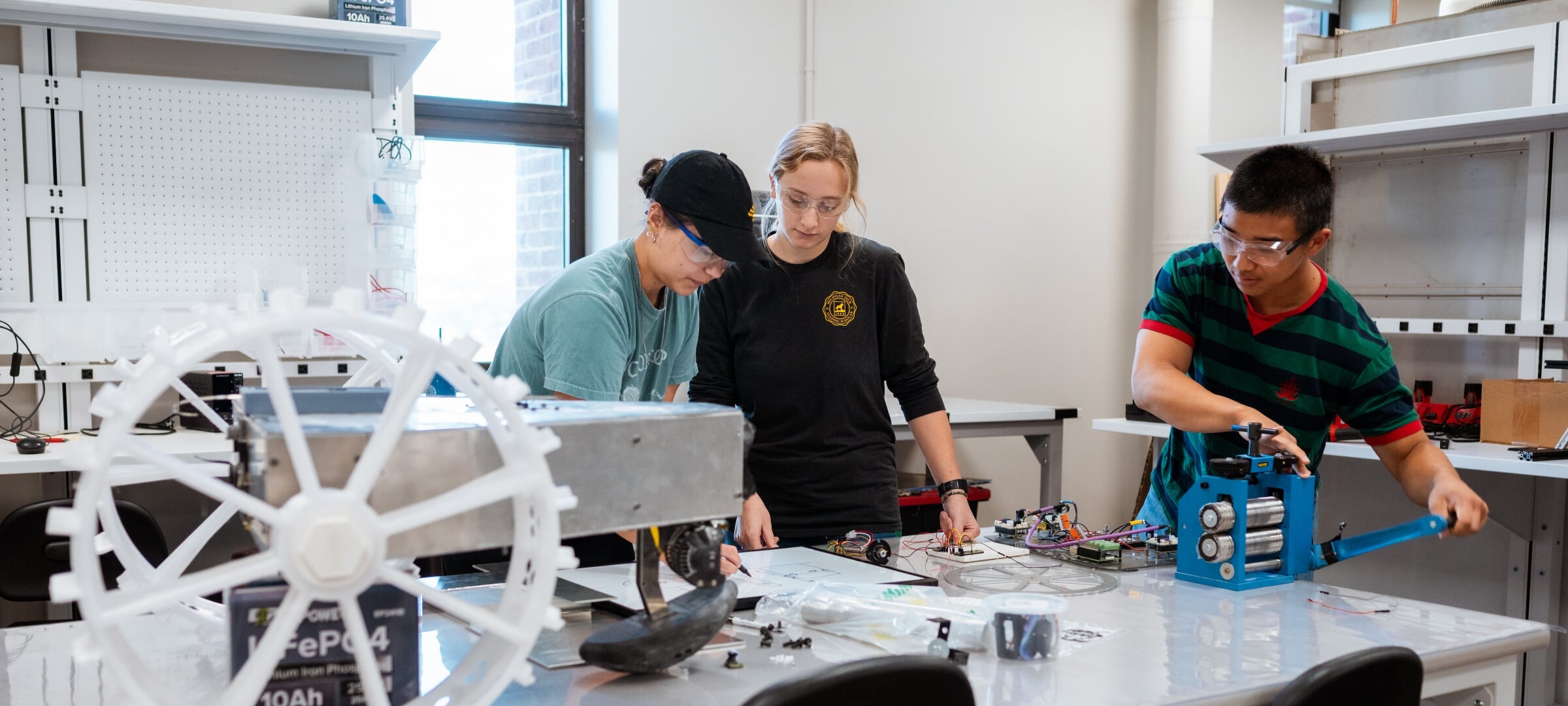 Three students in safety glasses work at a lab bench in a bright workshop.