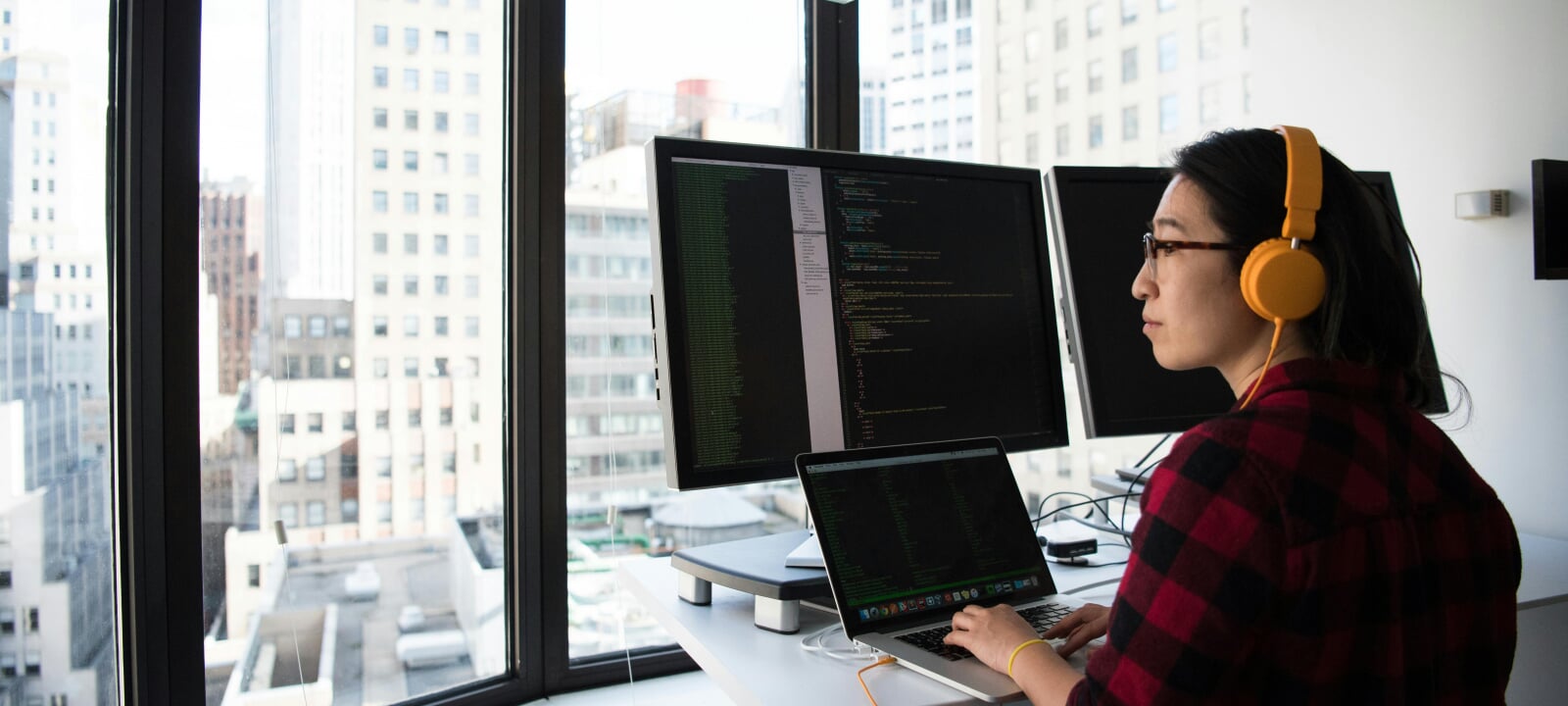 Young female engineer working on a computer while looking out a window onto an urban settting.