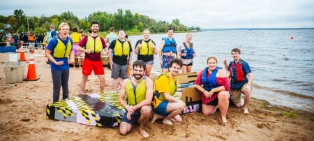 Ten Michigan Tech students pose on a beach with their cardboard boat, as other teams stage their boats in the background and race organizers survey the scene. There are sailboats moored in water in the background, along with a large orange buoy.