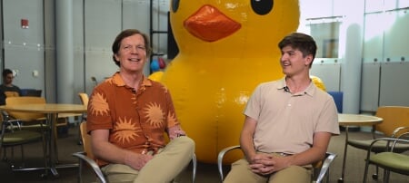 Professor Chuck Wallace and his student sit in a lounge with a large yellow rubber ducky in the background.