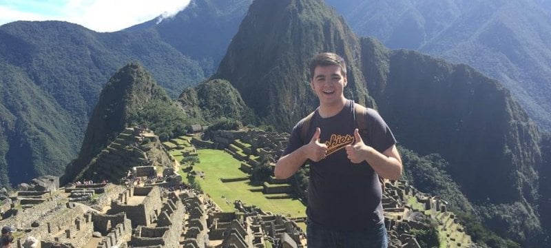 Student with two thumbs up standing in front of ruins in the mountains.