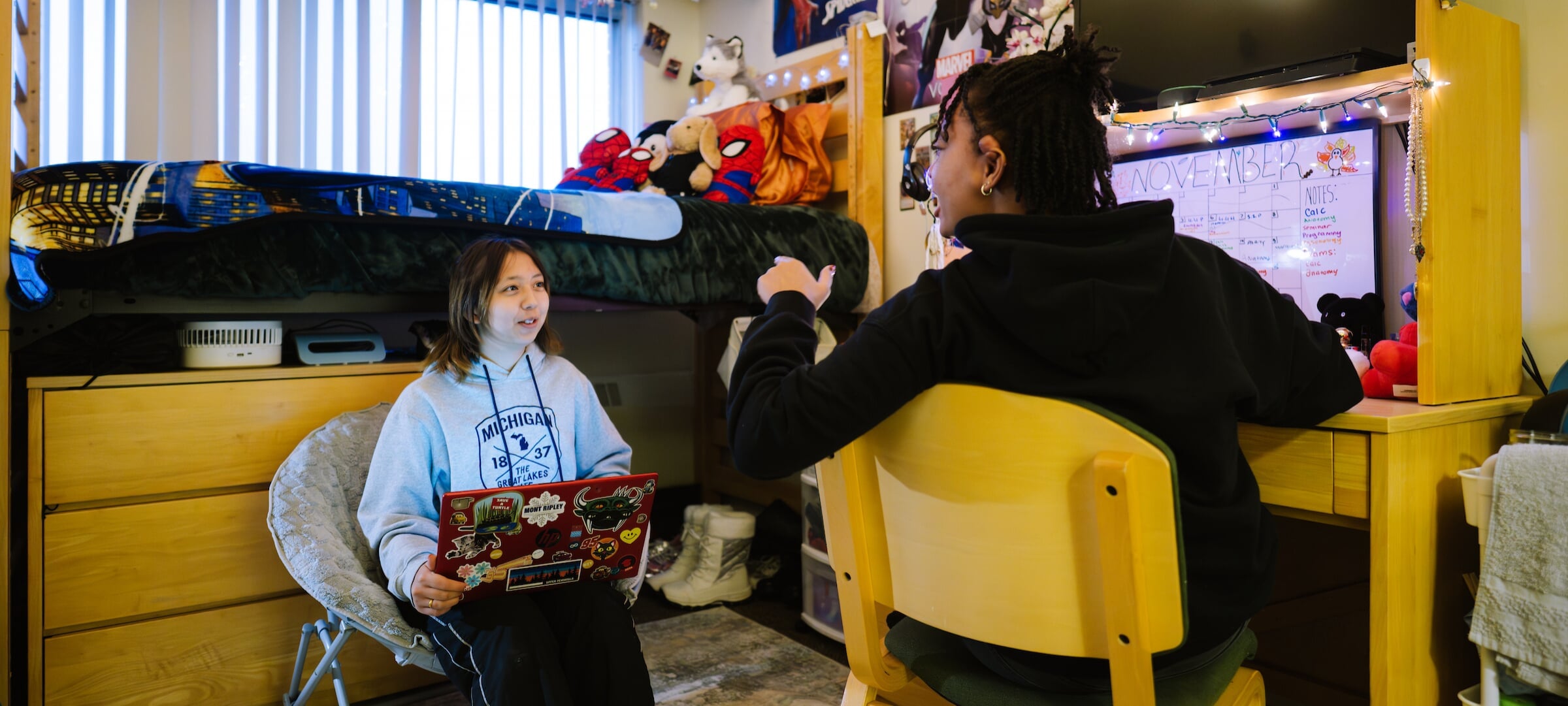 Two students in a dorm room talking with one sitting in a comfortable chair and the other at their desk