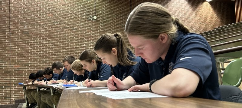 A large group of cadets sit inside the a building while taking a test.