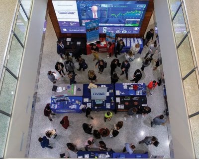 Looking down from above on several vendor tables and a giant stock market screen with people walking around.