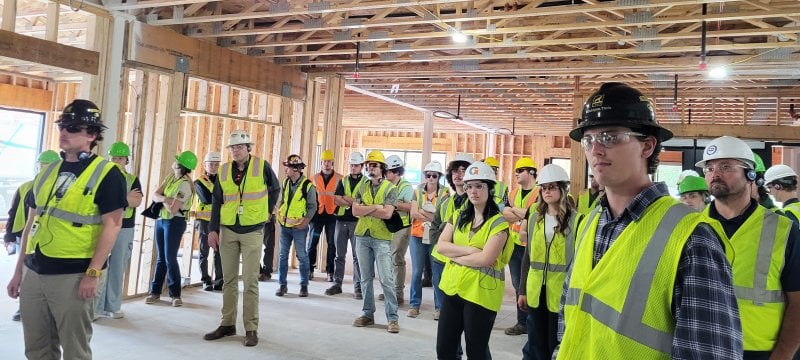 Students in hardhats and vests walking through a large room under construction with just studs and trusses visible..