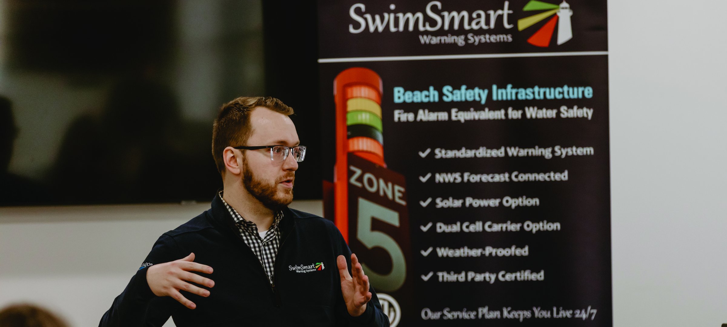 Jacob Soter standing in front of a sign explaining SwimSmart warning Systems Beach Safety Infrastructure and the options.