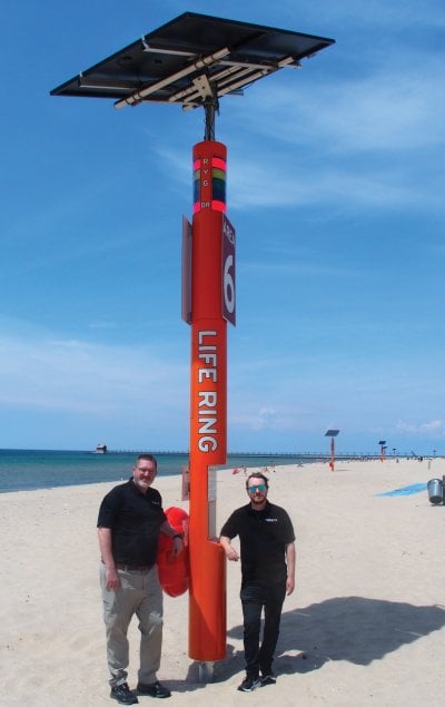 Two people standing next to a tower almost three times their height with a solar panel on top, the words Life Ring on the side, and a sign that says Area 6.