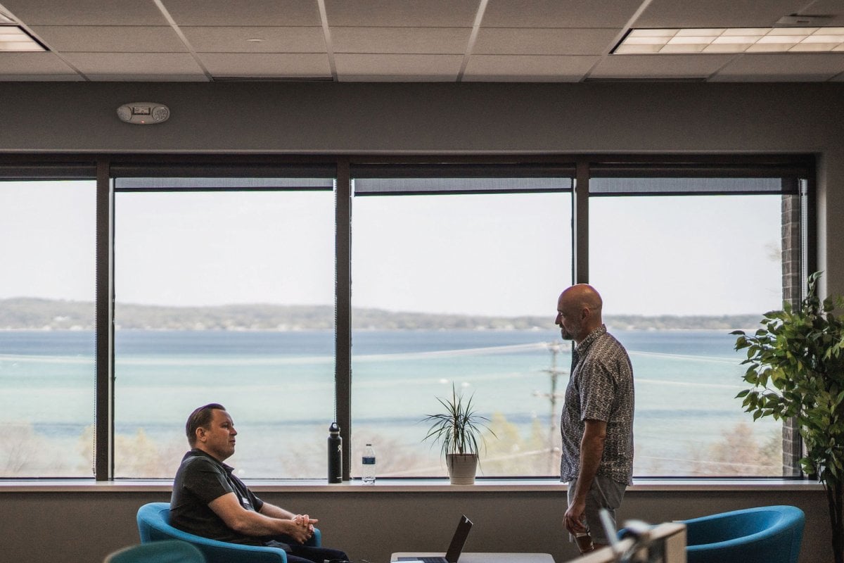 Two people, one sitting and one standing, talking in an open area in front of large windows.