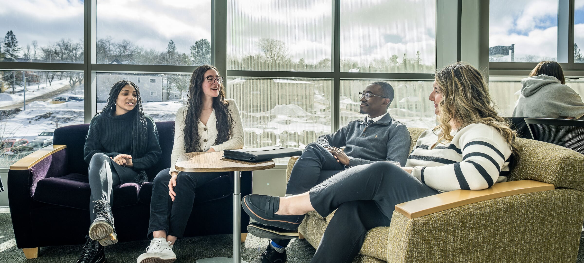 A group of College of Business students talking together in the library with a snowy campus behind them.