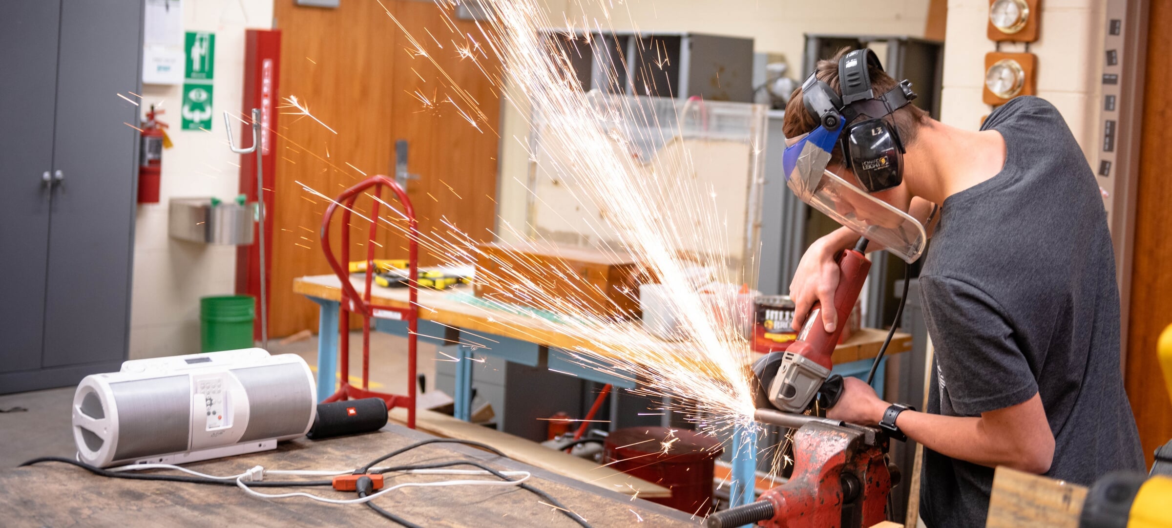 A student using a angle grinder on a pipe in a vice causing sparks to shoot out away from them.