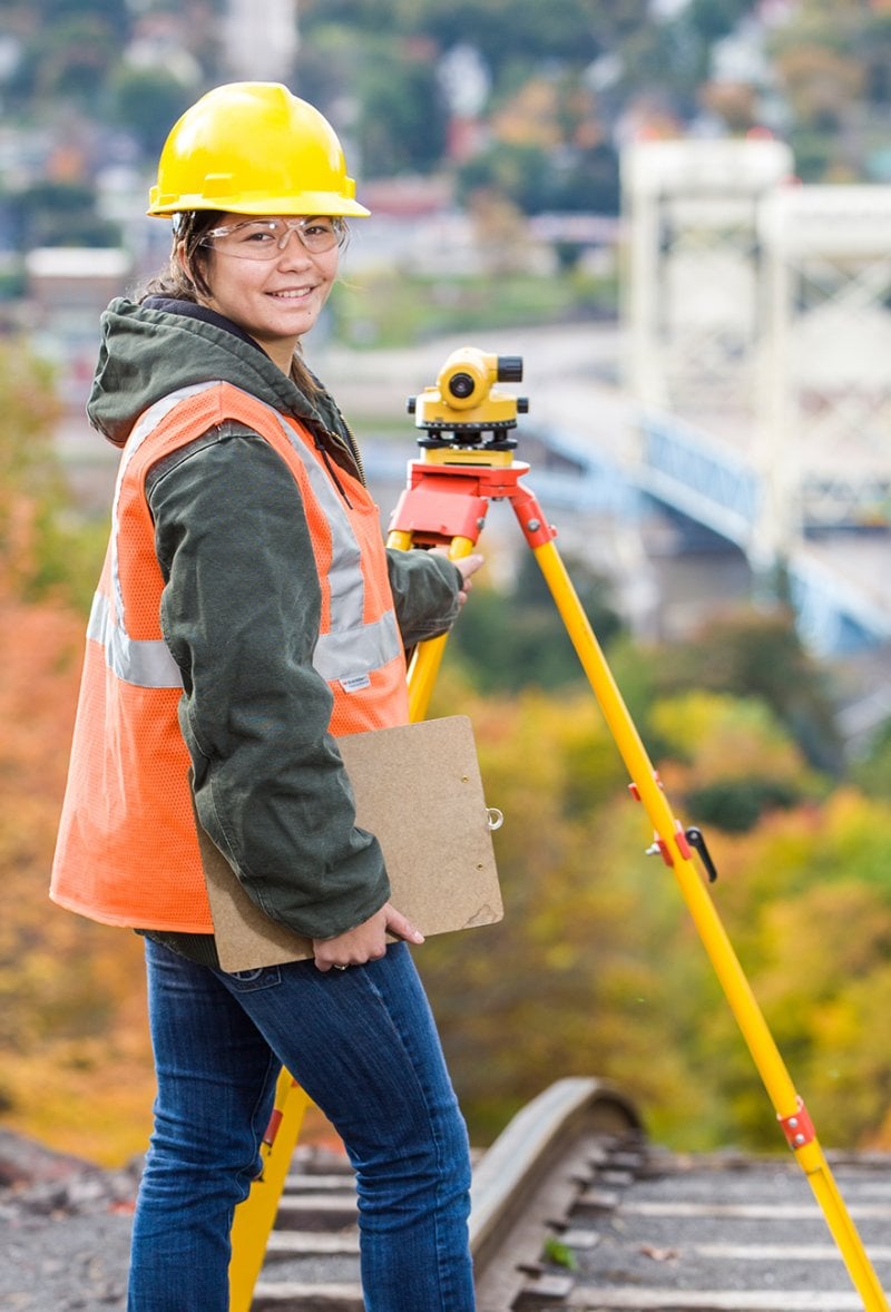 Student using surveying equipment with hard hat, vest, and cllpboard.