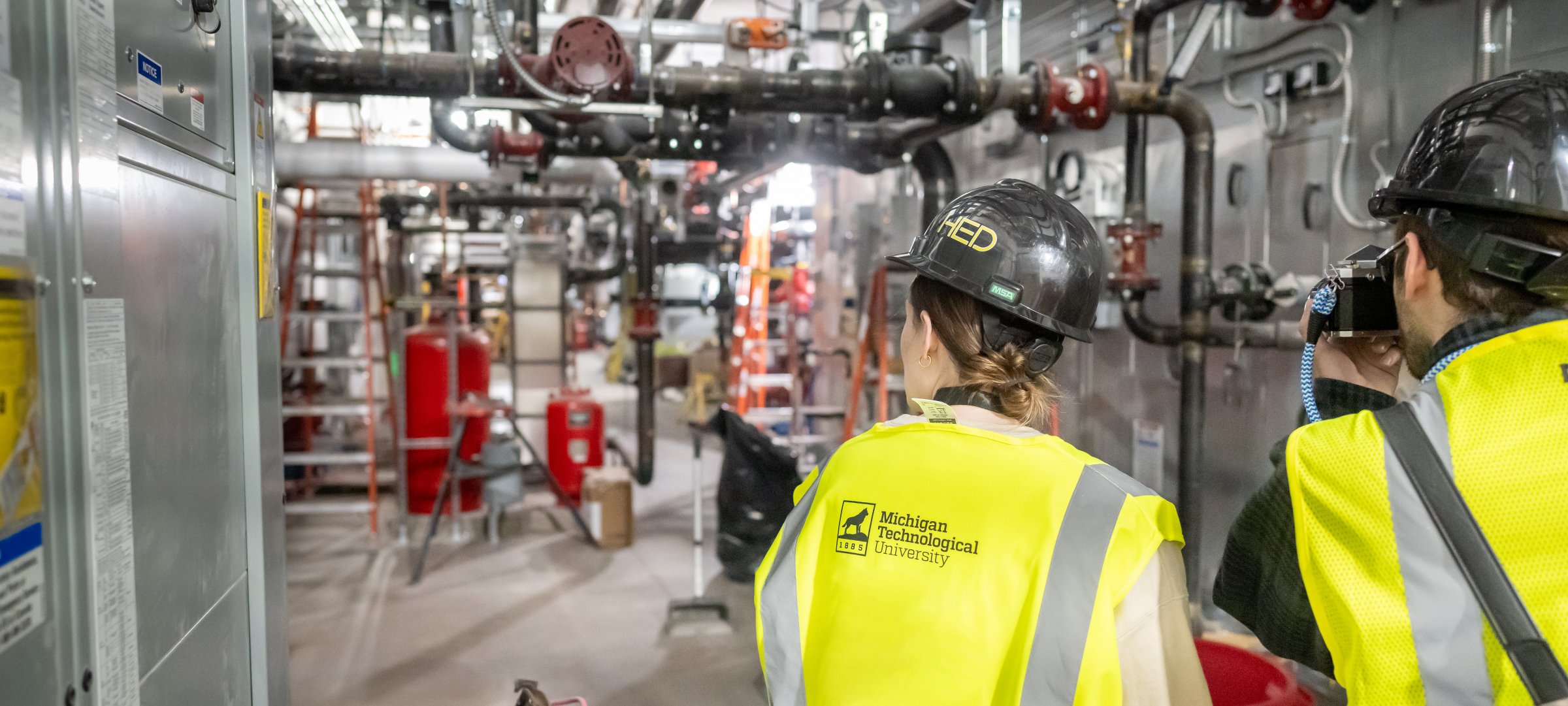 Two MTU civil students with hardhats and vests looking and photographing pipes inside the HSTEM building.