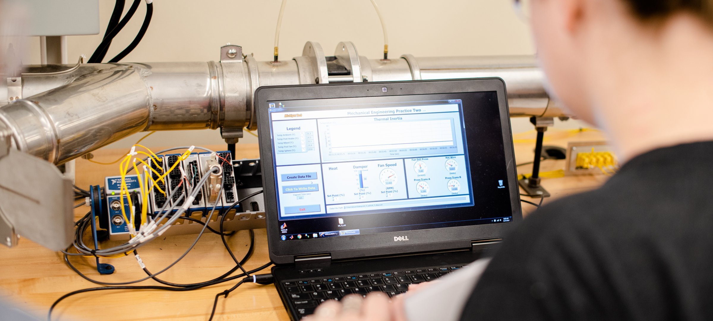 Person working at a laptop in front of some pipes and wires.