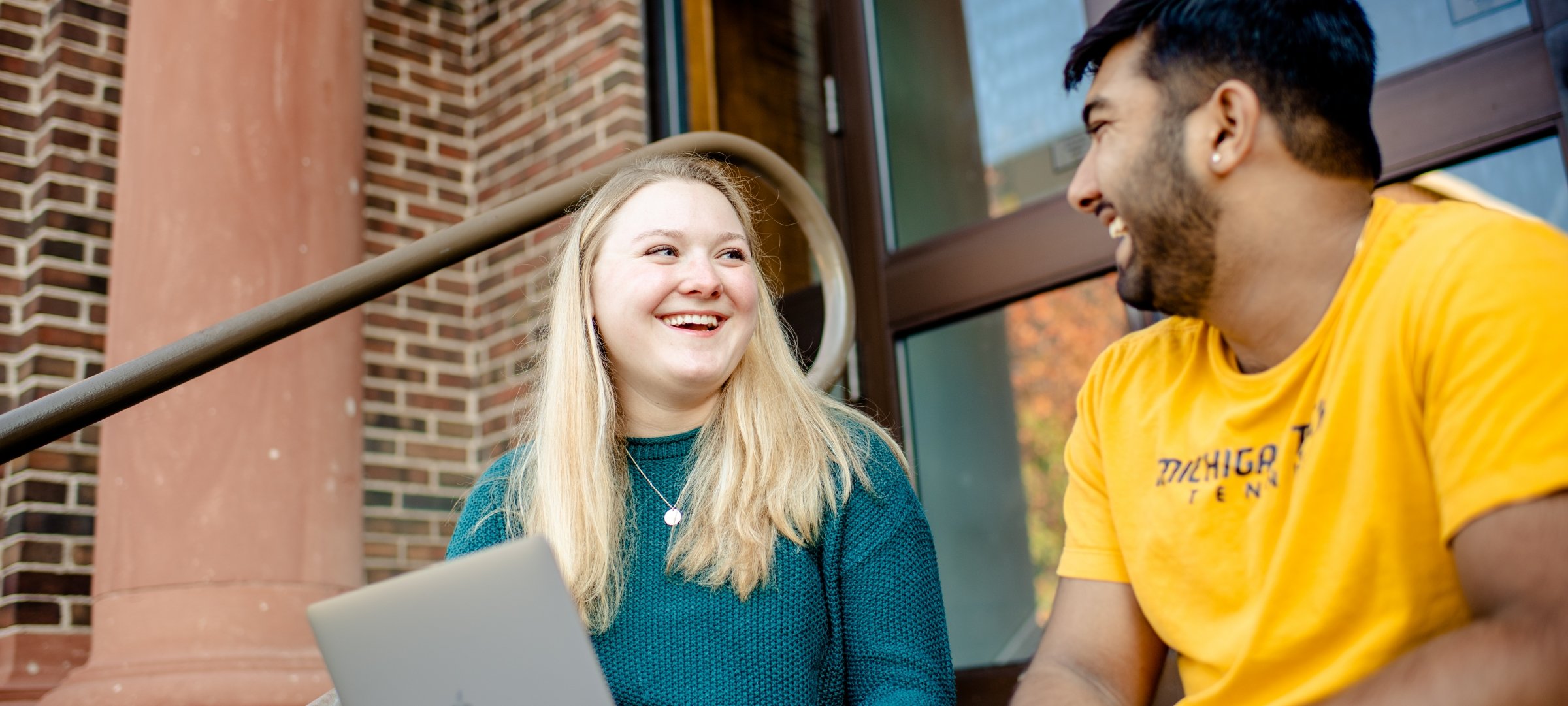 Two students smiling with a laptop on the front steps of an academic building