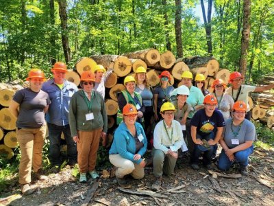 Sixteen people with hardhats in front of a log pile.