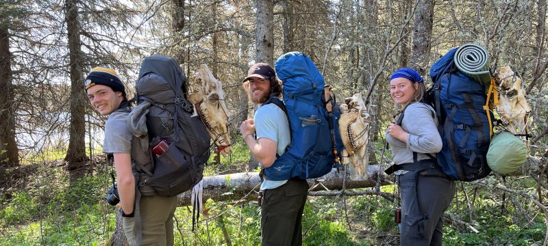 Three people in the woods with loaded hiking backpacks including skull bones attached to the outside.