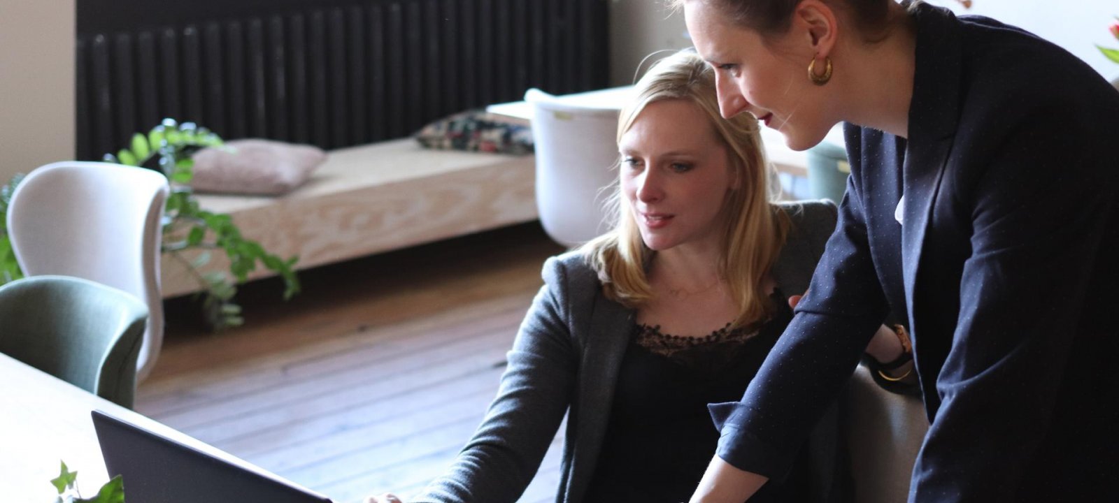 Two female professionals look at a computer. They are in an office setting