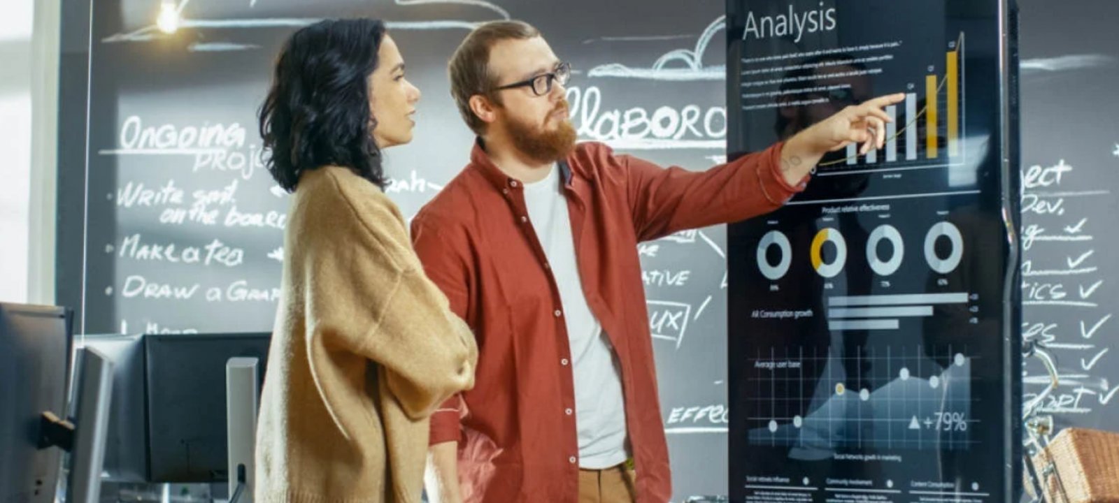 Two professionals analyze data while standing in front of a large monitor.