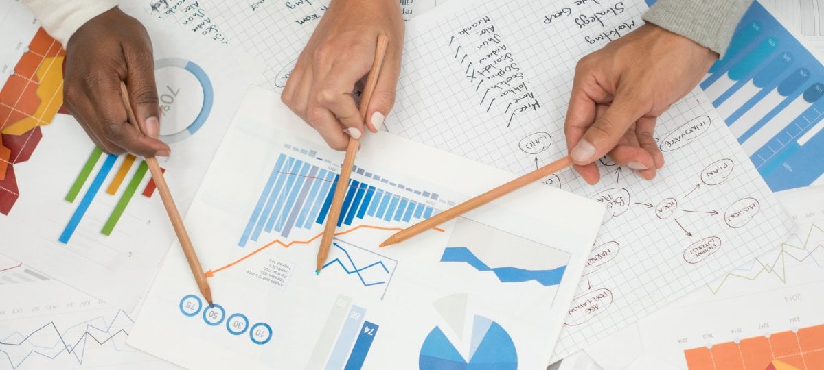 Three people, represented by hands with pencils, pointing at pages with statistical graphs on them.