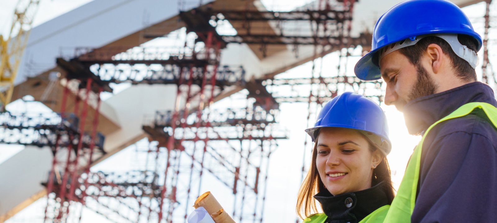 Two engineers, one male and one female, stand in front of a structure while looking at plans.