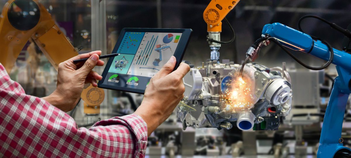 An engineer in a manufacturing setting with a lap top stands in front of a robotic machine, measuring its operation.