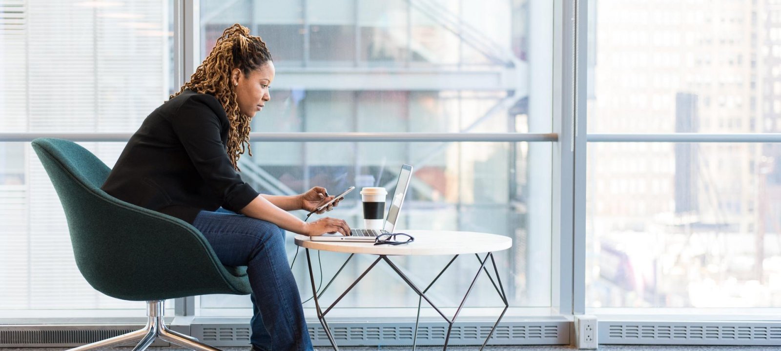 African American woman sitting in a teal chair while working on her computer and looking at her phone.