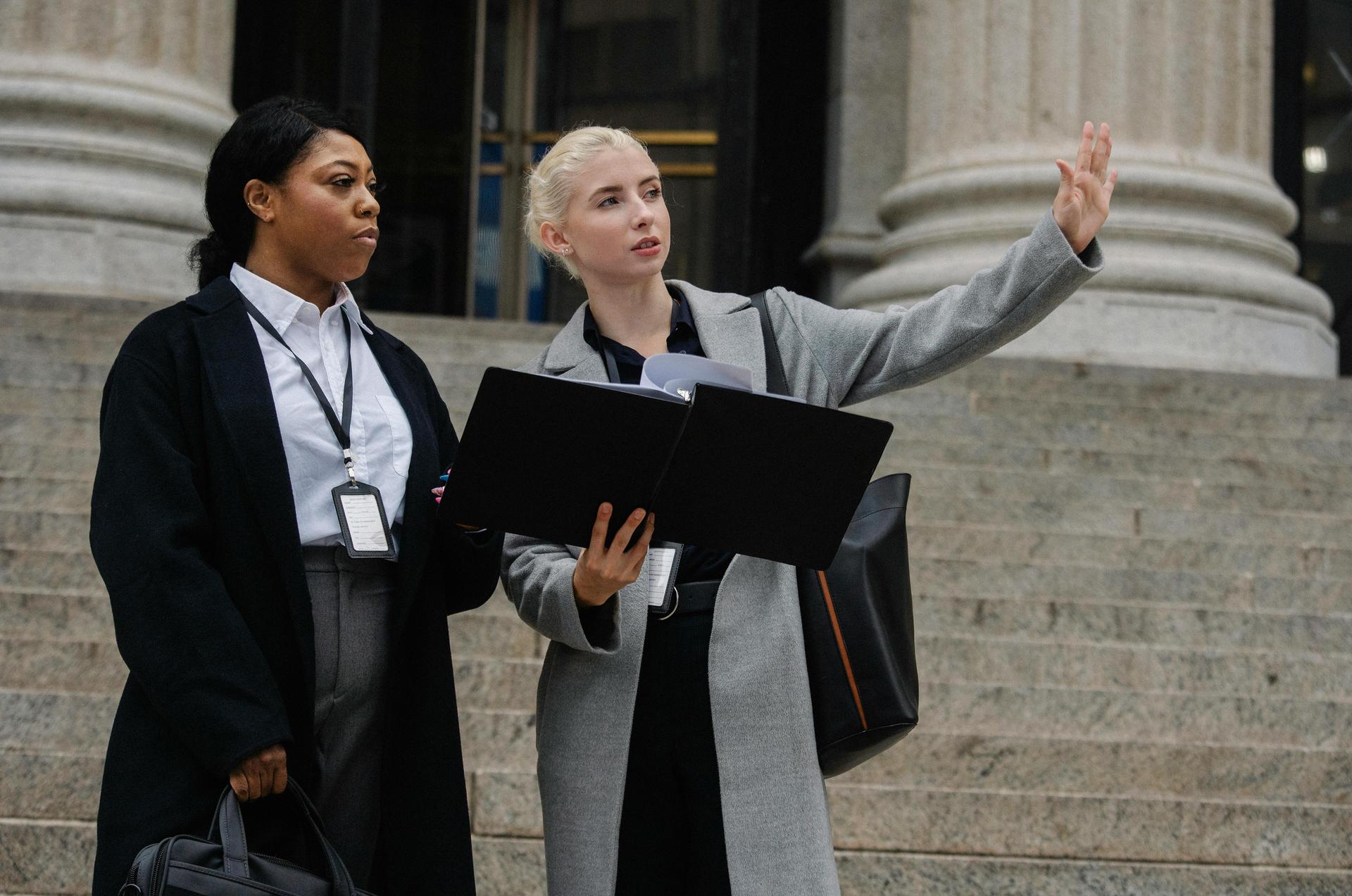Two female professionals discuss policy while standing on the steps of a government building.