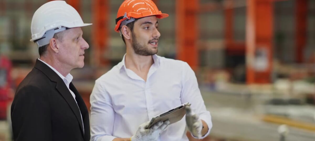 Two engineers in a factory setting discuss a plan. Both are wearing hard hats and one holds a metal clip board