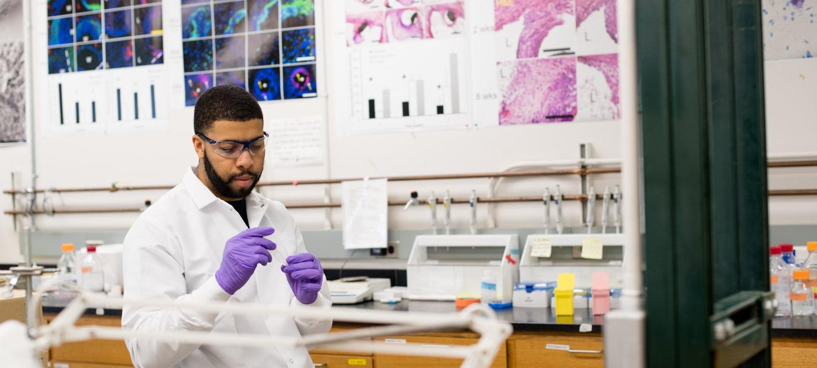 Person working in a lab, wearing a lab coat and gloves, surrounded by equipment