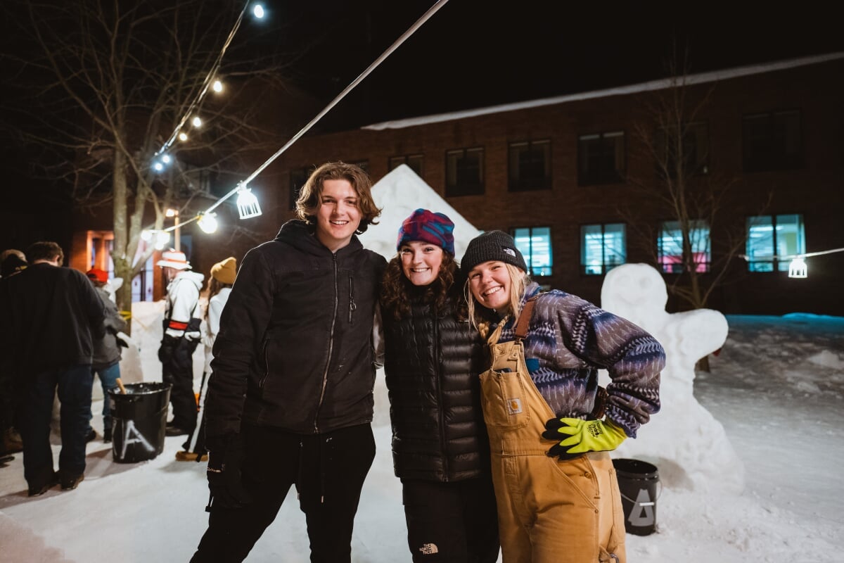 With more than 200 inches of annual snowfall, Houghton is one of the snowiest cities in the US. Three students smiling at the camera in front of a snow statue at night.