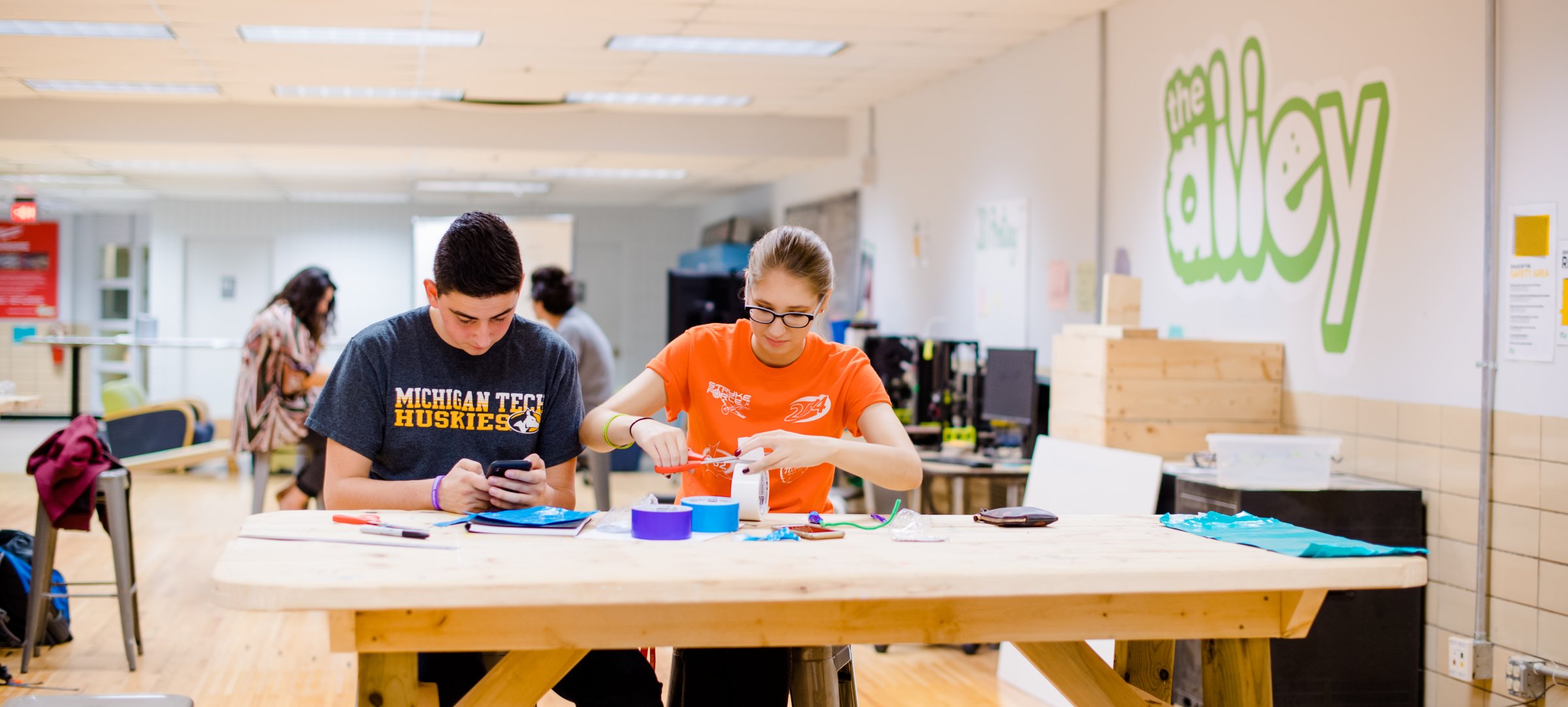 two students working at a tall wooden table covered with supplies with a wall to the right that says "the alley"