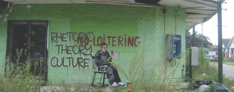 Rhetoric, Theory, Culture graphic on the side of a building with No Loitering spraypainted and the painted image of an elderly man sitting a rocking chair holding the U.S. flag