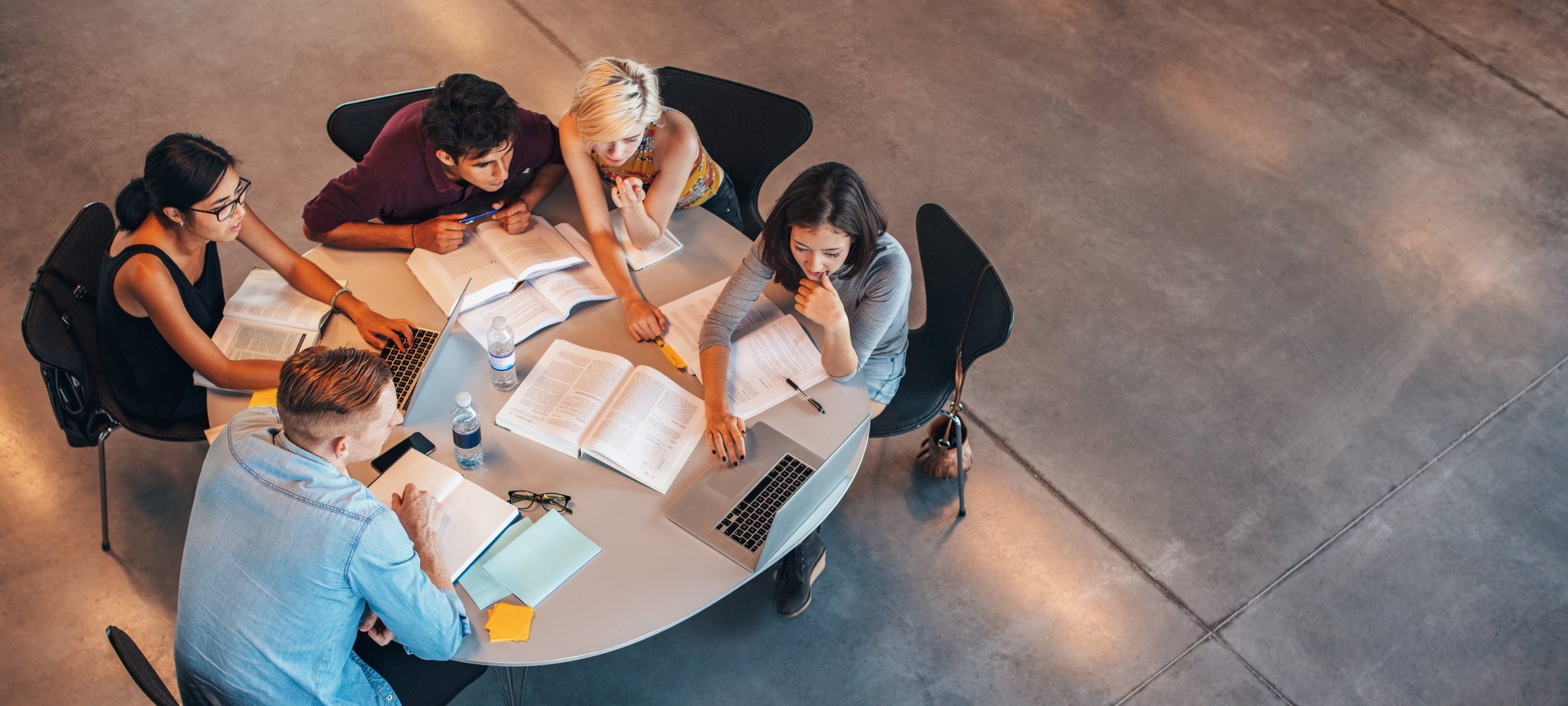 Five students sitting at a round table looking at a laptop with books open.