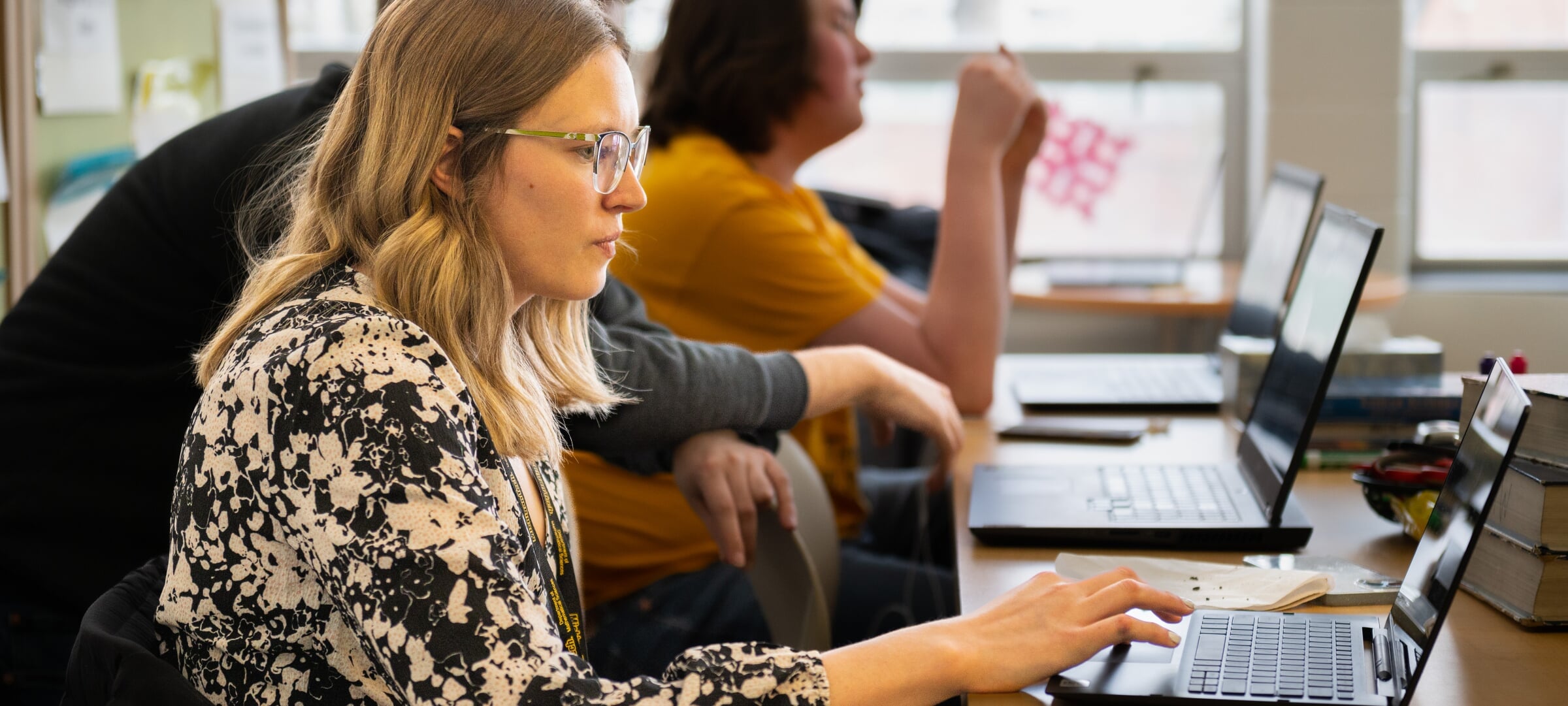 A woman looks intently at a laptop computer while others in the background work on their own laptops.