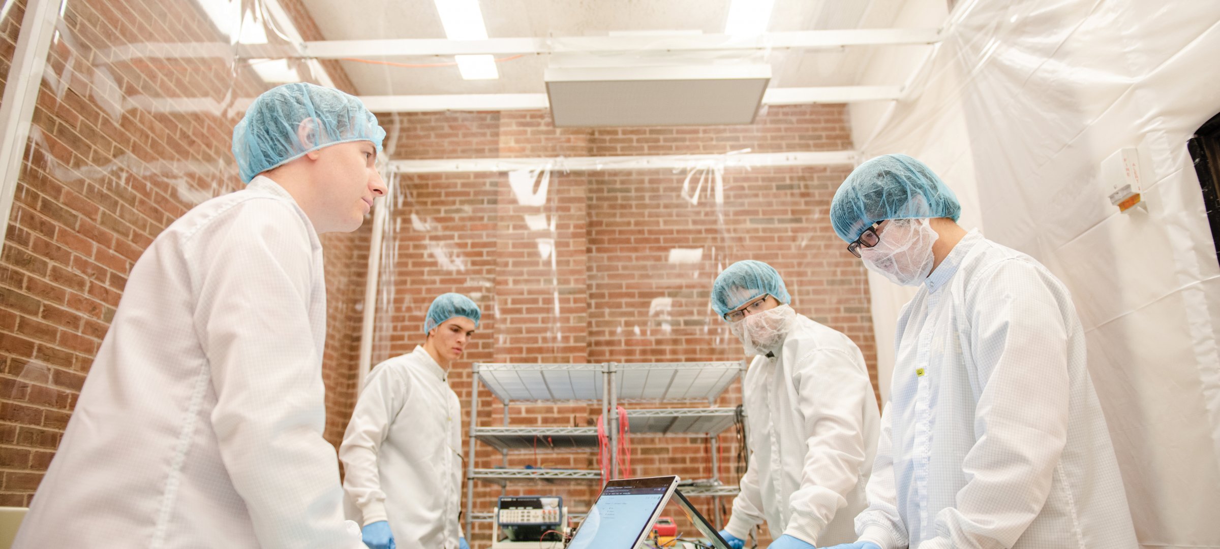 Four students wearing hairnets, coats, and gloves in a clean room at Michigan Technological University where they are building a nanosatellite.