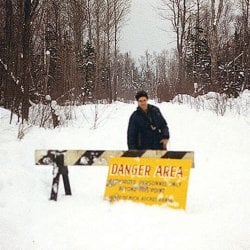 Man standing behind a baricade and a sign reading &quot;Danger Area Authorized Personnel Only Beyond This Point&quot; with additional words obscured by snow.