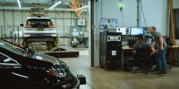 Two men look at computer screens in an automobile garage with two cars.