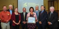 Eight staff members, board members and government officials stand in a ballroom smiling at the camera following the presentation of a framed award, held by a woman staff member in the center of the picture.