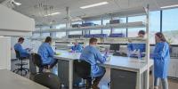 Six students in blue lab coats, purple gloves, and eye protection handle various medical equipment at a lab table.