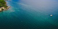 Drone aerial view of blue Lake Superior water on sunny day with small strip of green trees and shoreline on left and research boat in bottom right corner.