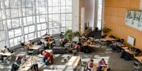 Aerial view of two dozen students studying at library tables with a wall of windows on left side letting in bright natural light.