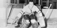 Black and white photo of dark-haired hockey goalie in Huskies sweater crouching with legs and arms out in front of a hockey goal, looking at the camera.