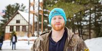 Owen Armstrong stands in front of the Michigan Tech clocktower wearing a camouflage jacket and a blue knit hat that reads â€œBlue Key Honor Society Winter Carnival Staff.â€