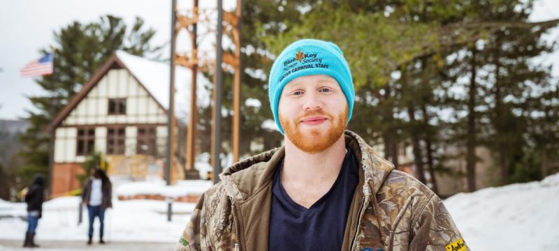 Owen Armstrong stands in front of the Michigan Tech clocktower wearing a camouflage jacket and a blue knit hat that reads â€œBlue Key Honor Society Winter Carnival Staff.â€